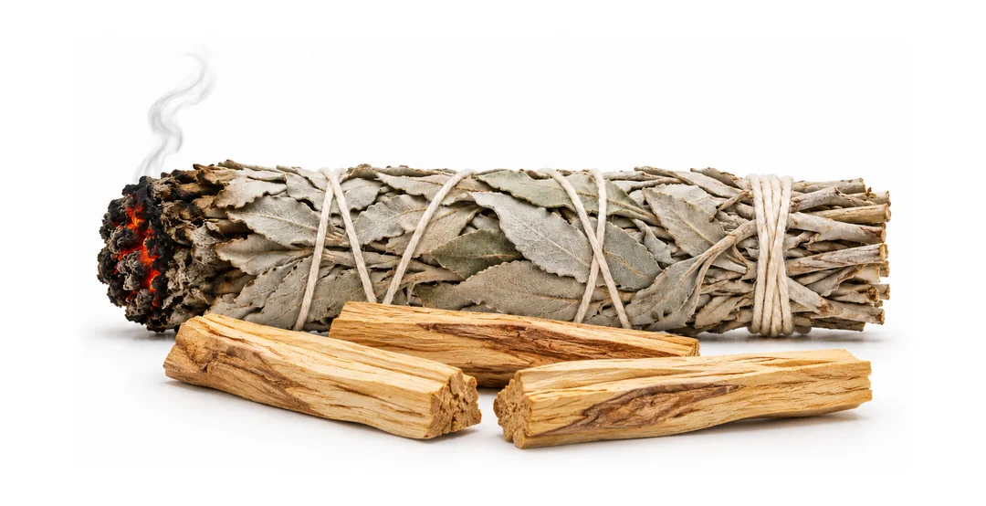 A smoldering white sage bundle beside three sticks of palo santo wood